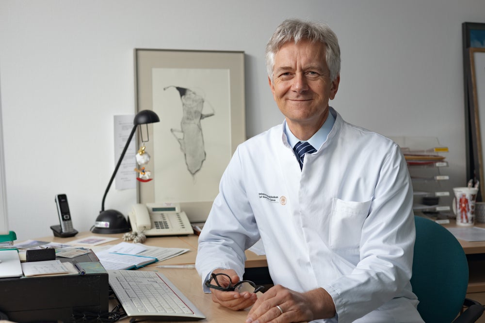 Prof. Dr. Johannes Schetelig wearing a white coat smiles gently at the camera and sitting at a desk in an office. He is holding glasses in his right hand, with papers, a keyboard, and a telephone visible on the desk behind him. Prof. Dr. Johannes Schetelig wearing a white coat smiles gently at the camera and sitting at a desk in an office. He is holding glasses in his right hand, with papers, a keyboard, and a telephone visible on the desk behind him.