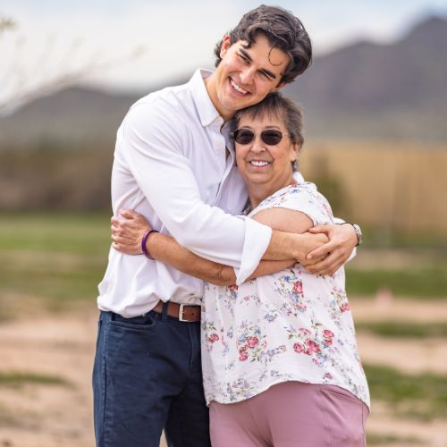 A young man and a woman, who are donor and patient, share a warm hug outdoors, both smiling joyfully. The man is wearing a white shirt and dark pants, while the woman is dressed in a floral blouse and pink pants. Mountains and a field can be seen in the blurred background. A young man and a woman, who are donor and patient, share a warm hug outdoors, both smiling joyfully. The man is wearing a white shirt and dark pants, while the woman is dressed in a floral blouse and pink pants. Mountains and a field can be seen in the blurred background.