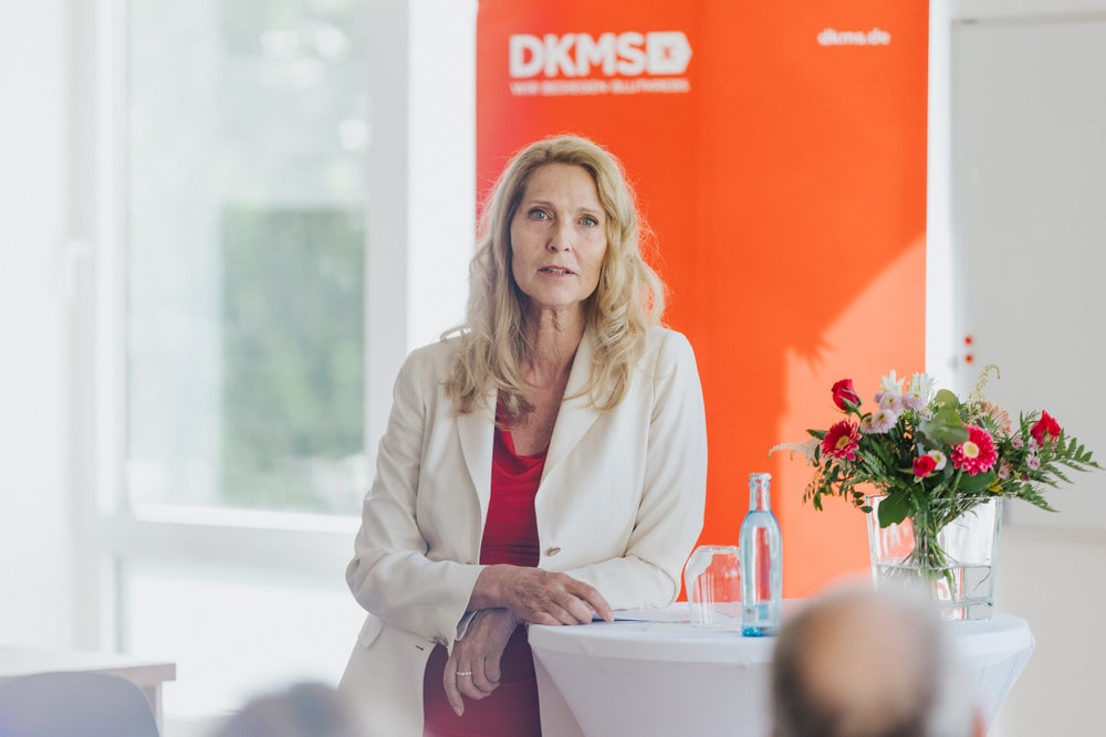 Dr. Elke Neujahr, Global CEO of the DKMS Group and Managing Director of the new DKMS Access To Treatment gGmbH, wearing a white blazer over a red top, speaks to an audience while standing next to a round table with a bouquet of flowers and a water bottle. An red DKMS banner is visible in the background. Dr. Elke Neujahr, Global CEO of the DKMS Group and Managing Director of the new DKMS Access To Treatment gGmbH, wearing a white blazer over a red top, speaks to an audience while standing next to a round table with a bouquet of flowers and a water bottle. An red DKMS banner is visible in the background.
