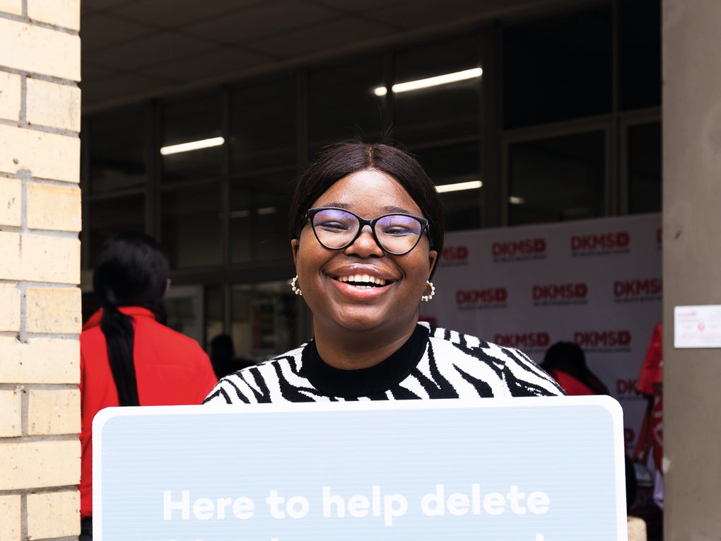 A black smiling woman wearing glasses and a black-and-white patterned sweater, holds a large light blue speech bubble sign that reads, "Here to help delete blood cancers and disorders". The DKMS logo is visible at the bottom of the sign. A black smiling woman wearing glasses and a black-and-white patterned sweater, holds a large light blue speech bubble sign that reads, "Here to help delete blood cancers and disorders". The DKMS logo is visible at the bottom of the sign.