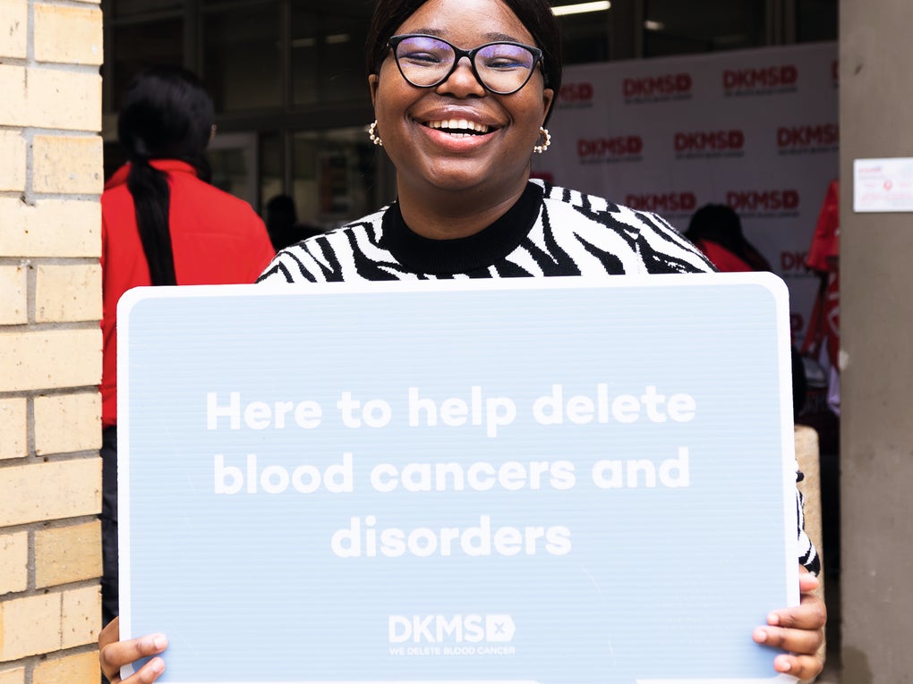 A black smiling woman wearing glasses and a black-and-white patterned sweater, holds a large light blue speech bubble sign that reads, "Here to help delete blood cancers and disorders". The DKMS logo is visible at the bottom of the sign. A black smiling woman wearing glasses and a black-and-white patterned sweater, holds a large light blue speech bubble sign that reads, "Here to help delete blood cancers and disorders". The DKMS logo is visible at the bottom of the sign.