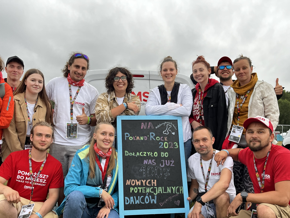 A group of thirteen enthusiastic DKMS volunteers pose together at the Poland Rock Festival 2023. They are standing and kneeling around a chalkboard that announces the number of new potential donors registered. The atmosphere is joyful and happy, with cloudy skies in the background. A group of thirteen enthusiastic DKMS volunteers pose together at the Poland Rock Festival 2023. They are standing and kneeling around a chalkboard that announces the number of new potential donors registered. The atmosphere is joyful and happy, with cloudy skies in the background.