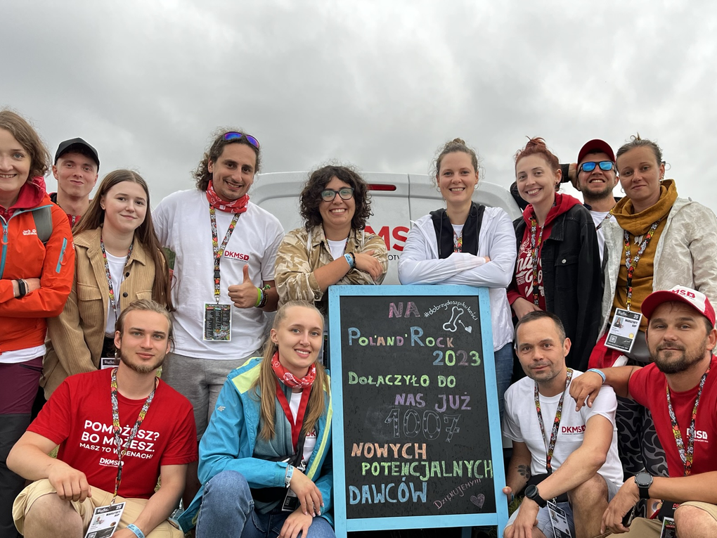 A group of thirteen enthusiastic DKMS volunteers pose together at the Poland Rock Festival 2023. They are standing and kneeling around a chalkboard that announces the number of new potential donors registered. The atmosphere is joyful and happy, with cloudy skies in the background. A group of thirteen enthusiastic DKMS volunteers pose together at the Poland Rock Festival 2023. They are standing and kneeling around a chalkboard that announces the number of new potential donors registered. The atmosphere is joyful and happy, with cloudy skies in the background.