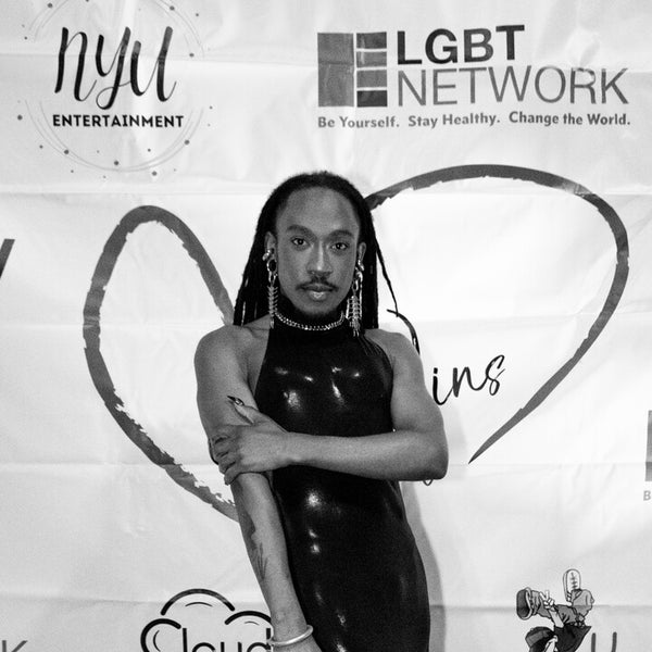 Black and white photo of the donor Deonté Lee posing confidently on a red carpet in front of a backdrop with various sponsor logos, including LGBT Network and NYU Entertainment. He's wearing a shiny black form-fitting, halter-neck gown, statement earrings, and has long dreadlocks. Black and white photo of the donor Deonté Lee posing confidently on a red carpet in front of a backdrop with various sponsor logos, including LGBT Network and NYU Entertainment. He's wearing a shiny black form-fitting, halter-neck gown, statement earrings, and has long dreadlocks.