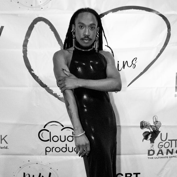 Black and white photo of the donor Deonté Lee posing confidently on a red carpet in front of a backdrop with various sponsor logos, including LGBT Network and NYU Entertainment. He's wearing a shiny black form-fitting, halter-neck gown, statement earrings, and has long dreadlocks. Black and white photo of the donor Deonté Lee posing confidently on a red carpet in front of a backdrop with various sponsor logos, including LGBT Network and NYU Entertainment. He's wearing a shiny black form-fitting, halter-neck gown, statement earrings, and has long dreadlocks.