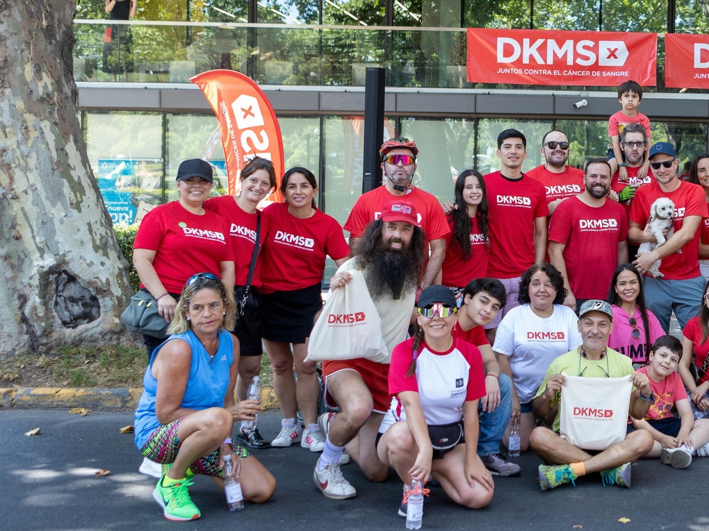 A large, cheerful group of DKMS volunteers, employess, supporters, and families pose together during a community event in Chile. Most are wearing red DKMS shirts and standing in front of a DKMS banner that reads 'Juntos contra el cáncer de sangre', which means 'Together against blood cancer'. A red sign on a bicycle trailer with giant cotton swabs and a QR code asks, '¿Cuántos kilómetros pedalearías para salvar una vida?', which means 'How many kilometers would you cycle to save a life?', encouraging donations and participation in a fundraising bike challenge. A large, cheerful group of DKMS volunteers, employess, supporters, and families pose together during a community event in Chile. Most are wearing red DKMS shirts and standing in front of a DKMS banner that reads 'Juntos contra el cáncer de sangre', which means 'Together against blood cancer'. A red sign on a bicycle trailer with giant cotton swabs and a QR code asks, '¿Cuántos kilómetros pedalearías para salvar una vida?', which means 'How many kilometers would you cycle to save a life?', encouraging donations and participation in a fundraising bike challenge.