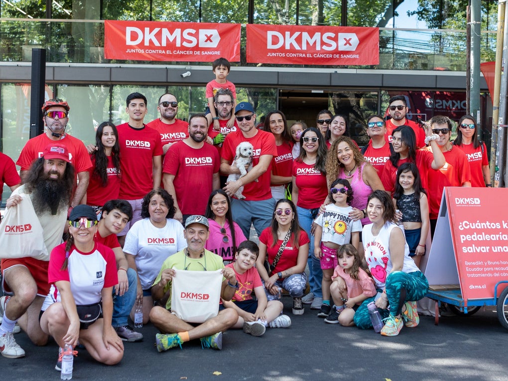 A large, cheerful group of DKMS volunteers, employess, supporters, and families pose together during a community event in Chile. Most are wearing red DKMS shirts and standing in front of a DKMS banner that reads 'Juntos contra el cáncer de sangre', which means 'Together against blood cancer'. A red sign on a bicycle trailer with giant cotton swabs and a QR code asks, '¿Cuántos kilómetros pedalearías para salvar una vida?', which means 'How many kilometers would you cycle to save a life?', encouraging donations and participation in a fundraising bike challenge. A large, cheerful group of DKMS volunteers, employess, supporters, and families pose together during a community event in Chile. Most are wearing red DKMS shirts and standing in front of a DKMS banner that reads 'Juntos contra el cáncer de sangre', which means 'Together against blood cancer'. A red sign on a bicycle trailer with giant cotton swabs and a QR code asks, '¿Cuántos kilómetros pedalearías para salvar una vida?', which means 'How many kilometers would you cycle to save a life?', encouraging donations and participation in a fundraising bike challenge.