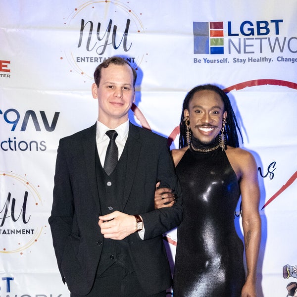 Two people posing together on a red carpet in front of banner. The man on the left is wearing a black suit and tie. The donor Deonté Lee on the right, wrapped his arm around the others man arm, is wearing a shiny black form-fitting, halter-neck gown with statement earrings and a confident smile. The background features logos from LGBT Network, NYU Entertainment, and other sponsors. Two people posing together on a red carpet in front of banner. The man on the left is wearing a black suit and tie. The donor Deonté Lee on the right, wrapped his arm around the others man arm, is wearing a shiny black form-fitting, halter-neck gown with statement earrings and a confident smile. The background features logos from LGBT Network, NYU Entertainment, and other sponsors.