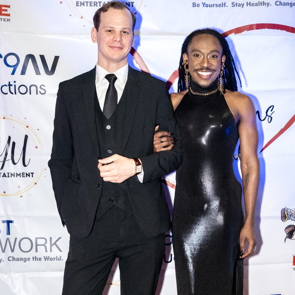 Two people posing together on a red carpet in front of banner. The man on the left is wearing a black suit and tie. The donor Deonté Lee on the right, wrapped his arm around the others man arm, is wearing a shiny black form-fitting, halter-neck gown with statement earrings and a confident smile. The background features logos from LGBT Network, NYU Entertainment, and other sponsors. Two people posing together on a red carpet in front of banner. The man on the left is wearing a black suit and tie. The donor Deonté Lee on the right, wrapped his arm around the others man arm, is wearing a shiny black form-fitting, halter-neck gown with statement earrings and a confident smile. The background features logos from LGBT Network, NYU Entertainment, and other sponsors.