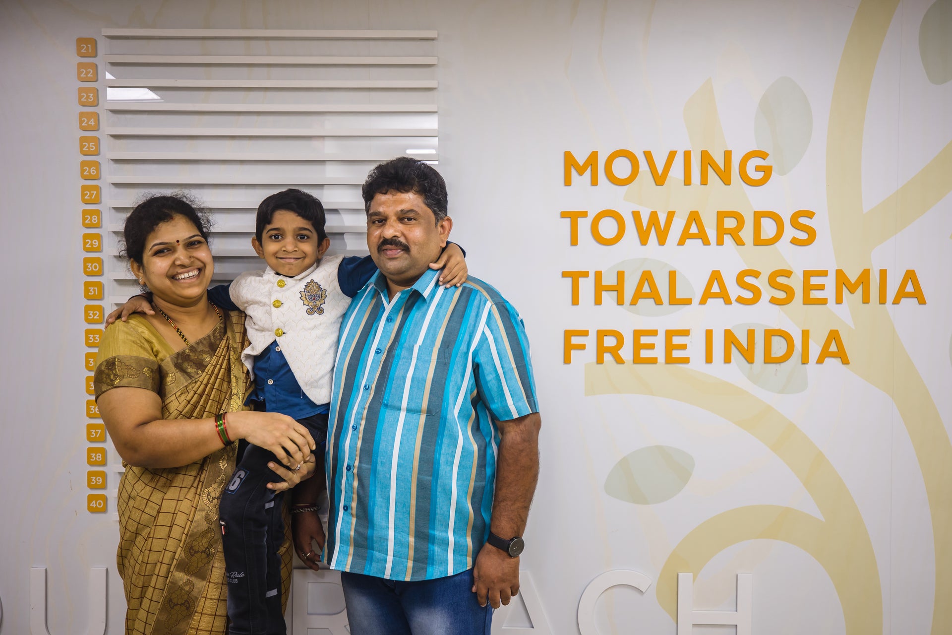 A smiling family of three stands in front of a wall that reads 'Moving Towards Thalassemia Free India.' The mother on the left, wearing a gold sari, holds their young son in her arms, while the father on the right, stands beside them in a blue striped shirt. All three are looking happily at the camera. A smiling family of three stands in front of a wall that reads 'Moving Towards Thalassemia Free India.' The mother on the left, wearing a gold sari, holds their young son in her arms, while the father on the right, stands beside them in a blue striped shirt. All three are looking happily at the camera.
