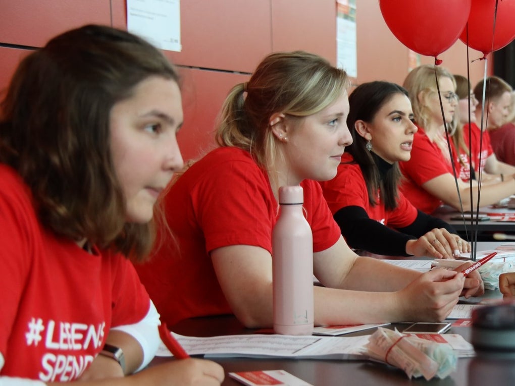 Young DKMS volunteers in red shirts sit at a long table on the left during a university registration event talking to students, which are sitting on the right, about registering as potential donors with DKMS. Swab kits and paperwork are on the table. Red balloons float above the tables, and the atmosphere is focused and energetic as people engage in conversation. Young DKMS volunteers in red shirts sit at a long table on the left during a university registration event talking to students, which are sitting on the right, about registering as potential donors with DKMS. Swab kits and paperwork are on the table. Red balloons float above the tables, and the atmosphere is focused and energetic as people engage in conversation.