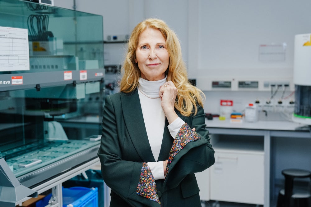 Dr. Elke Neujahr, Global CEO of DKMS Group, stands in a modern laboratory. She wears a black blazer with floral-patterned sleeve linings and a white turtleneck, looking directly into the camera with a gentle smile. Scientific equipment and lab machinery are visible in the background. Dr. Elke Neujahr, Global CEO of DKMS Group, stands in a modern laboratory. She wears a black blazer with floral-patterned sleeve linings and a white turtleneck, looking directly into the camera with a gentle smile. Scientific equipment and lab machinery are visible in the background.