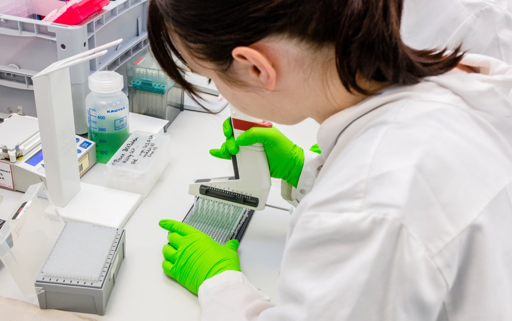 A female scientist with a brown ponytail shown from behind, wearing a white lab coat and bright green protective gloves uses a pipette-device with eight pipettes to fill a multichannel plate in a laboratory. Various lab supplies and containers are visible on the white table around her. A female scientist with a brown ponytail shown from behind, wearing a white lab coat and bright green protective gloves uses a pipette-device with eight pipettes to fill a multichannel plate in a laboratory. Various lab supplies and containers are visible on the white table around her.