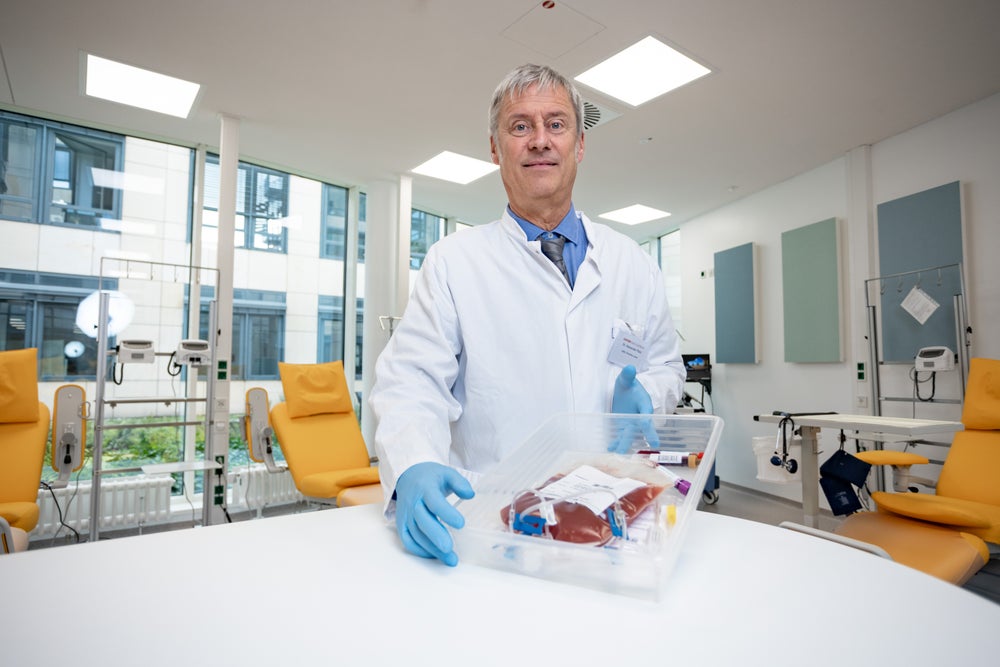 The Medical Director of the DKMS Stem Cell Bank Dr. Alexander Platz, wearing a white lab coat, blue gloves, and a tie, is holding an Adult Donor Cryopreserved Unit (ADCU) in a transparent container above a white table. He is standing in a modern clinical room with yellow chairs. The Medical Director of the DKMS Stem Cell Bank Dr. Alexander Platz, wearing a white lab coat, blue gloves, and a tie, is holding an Adult Donor Cryopreserved Unit (ADCU) in a transparent container above a white table. He is standing in a modern clinical room with yellow chairs.