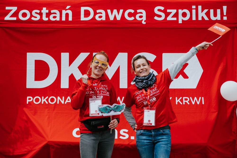 Two smiling female DKMS volunteers stand in front of a red DKMS booth with the Polish message 'Zostań Dawcą Szpiku!', which means 'Become a stem cell donor!'. They are wearing red DKMS shirts, event badges. The woman, who stands on the right, holds a small DKMS flag and the other woman, who stands on the left, has two funny signs with a pair of glasses and with wings in both hands. The atmosphere is joyful. Two smiling female DKMS volunteers stand in front of a red DKMS booth with the Polish message 'Zostań Dawcą Szpiku!', which means 'Become a stem cell donor!'. They are wearing red DKMS shirts, event badges. The woman, who stands on the right, holds a small DKMS flag and the other woman, who stands on the left, has two funny signs with a pair of glasses and with wings in both hands. The atmosphere is joyful.