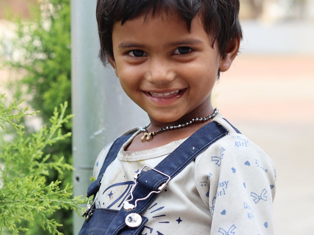 A young boy with black hair smiles warmly at the camera while standing outdoors next to green plants. The boy is wearing a light-colored shirt with printed patterns and denim overalls, and a beaded necklace. The background is softly blurred. A young boy with black hair smiles warmly at the camera while standing outdoors next to green plants. The boy is wearing a light-colored shirt with printed patterns and denim overalls, and a beaded necklace. The background is softly blurred.