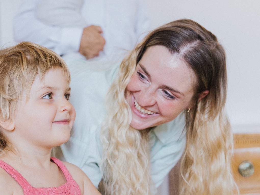 A smiling woman with long blond hair is bending down and looking at her little daughter with blond hair and a pink dress. The girl is smiling softly and looking slightly to the right side. The mood is warm and joyful, with other people blurred in the background. A smiling woman with long blond hair is bending down and looking at her little daughter with blond hair and a pink dress. The girl is smiling softly and looking slightly to the right side. The mood is warm and joyful, with other people blurred in the background.