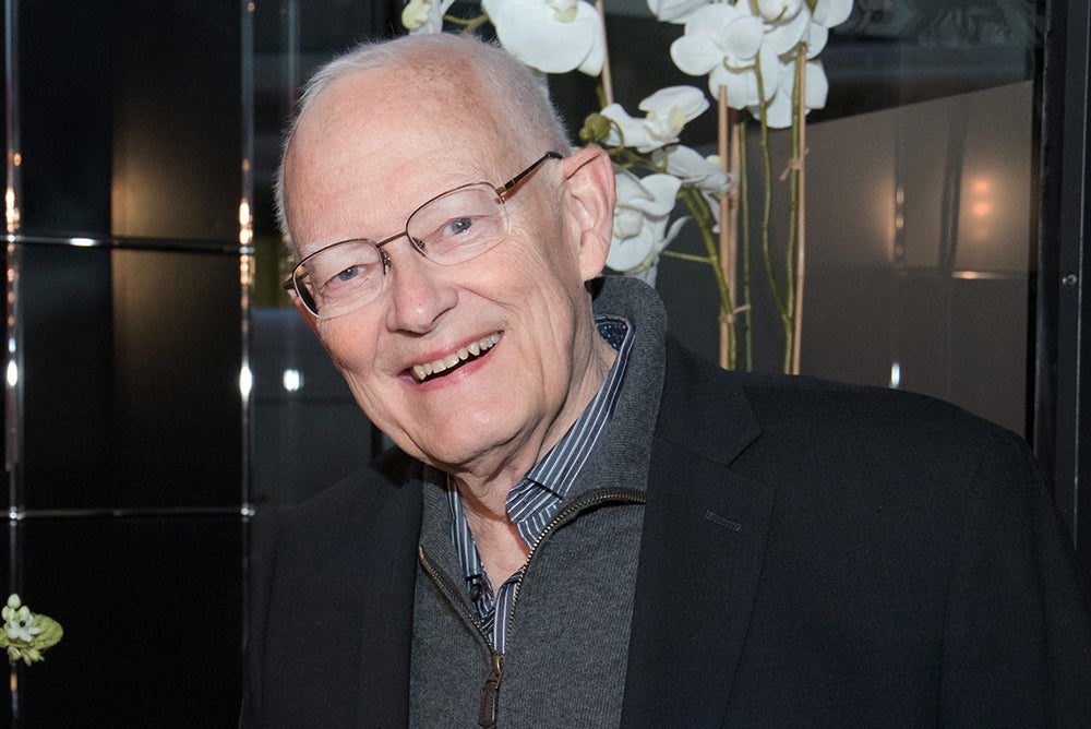 John Hansen, wearing glasses, a dark blazer, a zip-up sweater, and a striped shirt stands in front of orchids at an indoor event. John Hansen, wearing glasses, a dark blazer, a zip-up sweater, and a striped shirt stands in front of orchids at an indoor event.