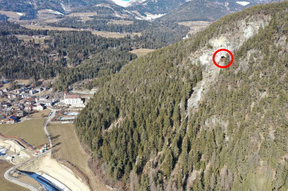 Il fianco del versante a rischio di caduta massi (il Koflloch è cerchiato in rosso) con il cantiere della circonvallazione di Chienes e la barriera provvisoria di protezione dalla caduta massi già eretta durante la fase di costruzione. (Immagine da drone: Ufficio tecnico strade Nord-Est)