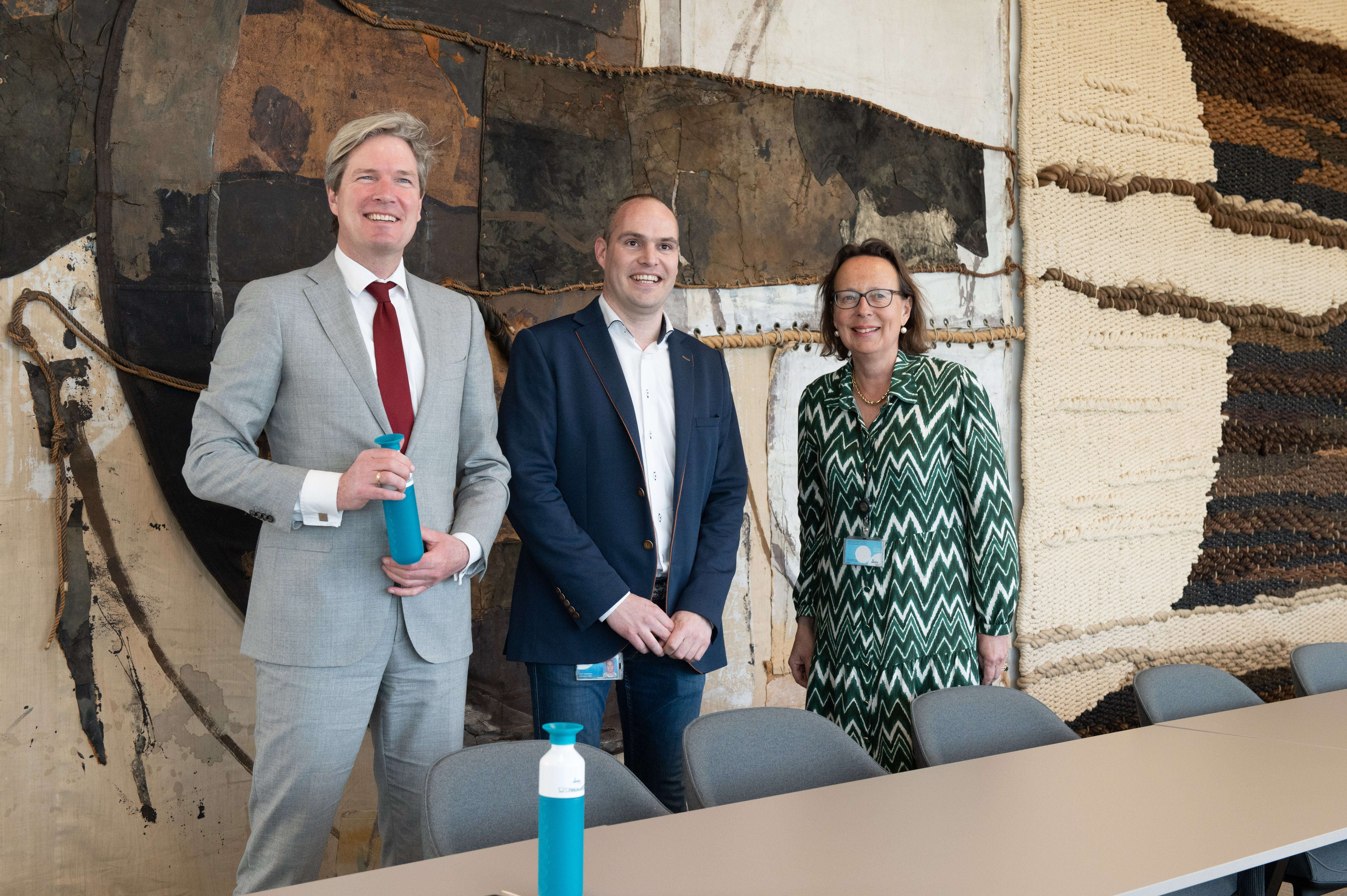 Foort van Oosten, burgemeester van Nissewaard, Paul van Versendaal, regiohoofd Groot Berenplaat en onze Algemeen Directeur Annette Ottolini poseren voor het wandkleed in een vergaderzaal van het hoofdgebouw op de Berenplaat.