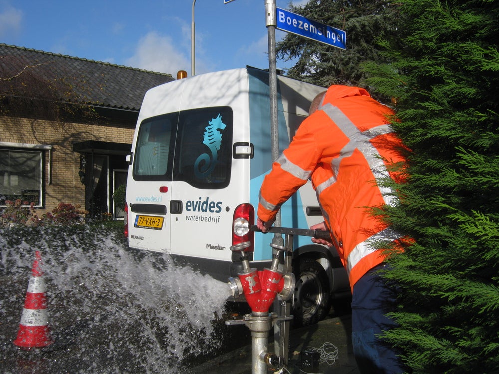 Een medewerker van Evides draait een waterkraan open. Het water spuit uit de kraan. Op de achtergrond staat een Evides bus.