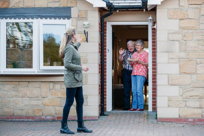 A woman waving to an older couple in the doorway of their home