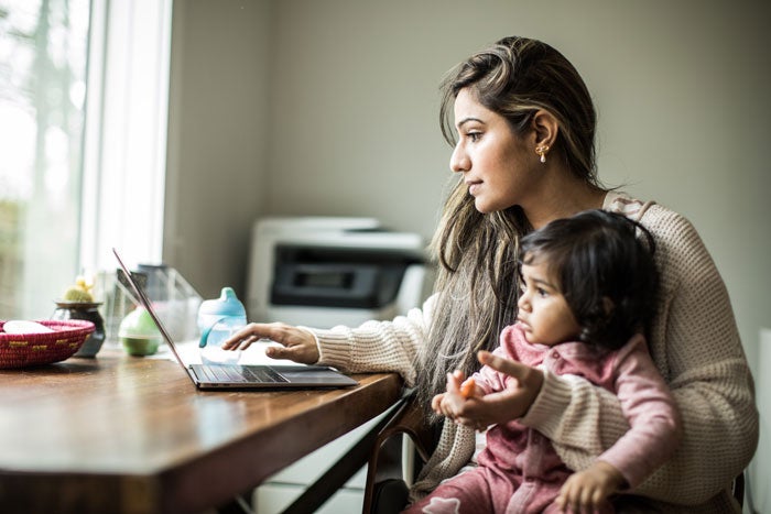A woman working at a laptop while holding a child on her lap