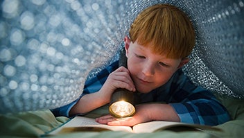 Young boy under a blanket using a torch to read a book