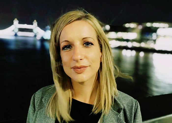 Sarah Andrews standing on a bridge, in London, at night, looking at the camera 