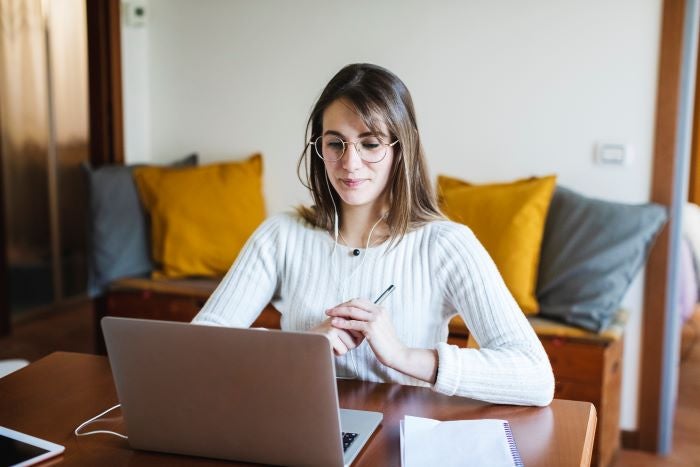 A lady sitting at a desk with a pen in her hand, looking at a laptop.