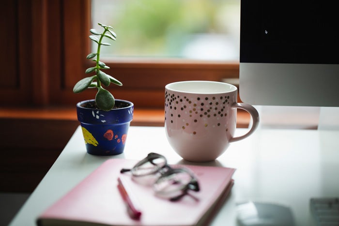 A close up image of a tea cup, small plant pot, notepad, pen and glasses sitting on a desk