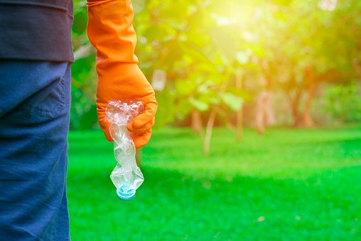 Close up of a person wearing a rubber glove and holding a plastic bottle in the foreground with grass and trees in the background