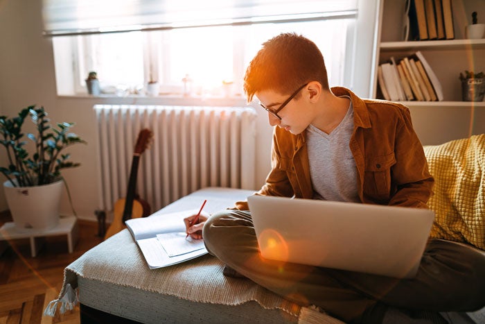 A young boy studying on the sofa with a laptop and open exercise book