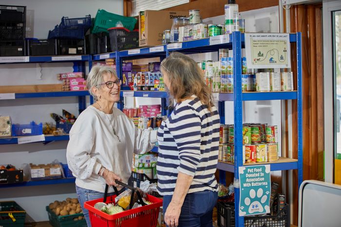 Two women talking and collecting items in a foodbank; one is holding a basket of food