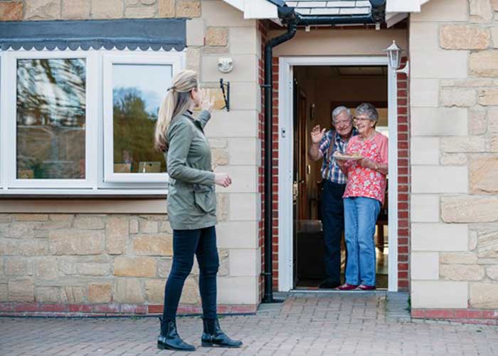 Elderly couple standing at front door waving to a younger woman