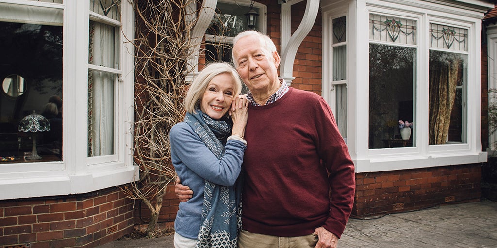 Joyful looking elderly couple standing outside their home whilst lovingly holding one another
