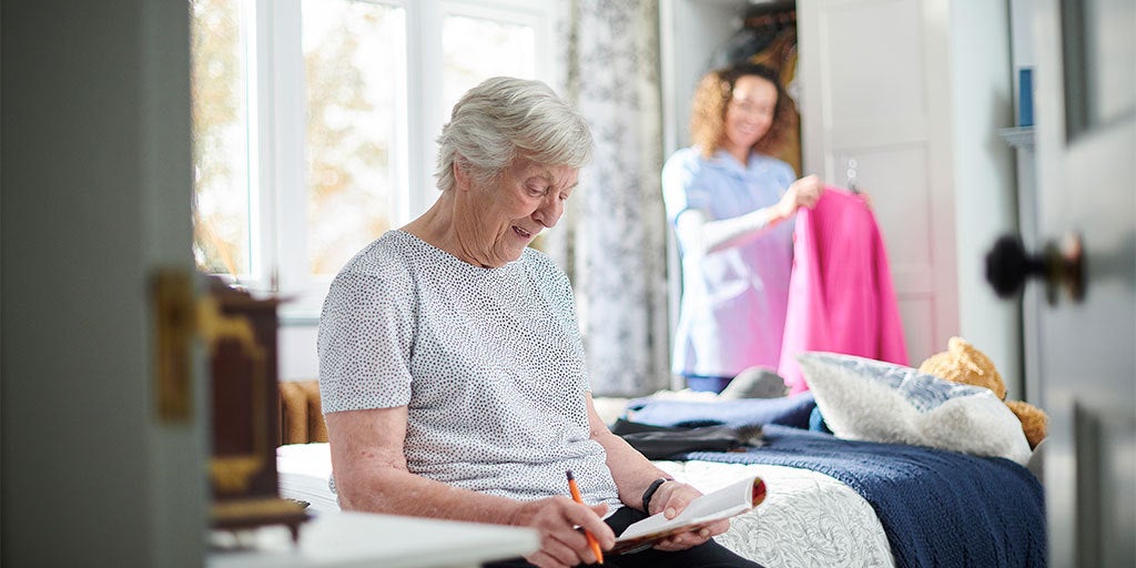 Elderly woman sitting on edge of the bed with notebook as nurse puts clothes away in the background