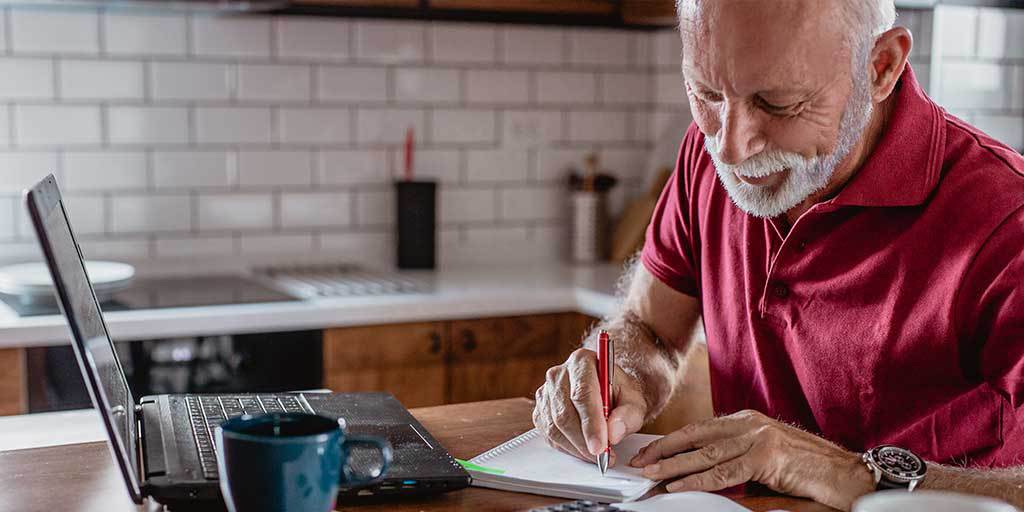 Elderly man sitting at kitchen table with laptop and writing on a notebook