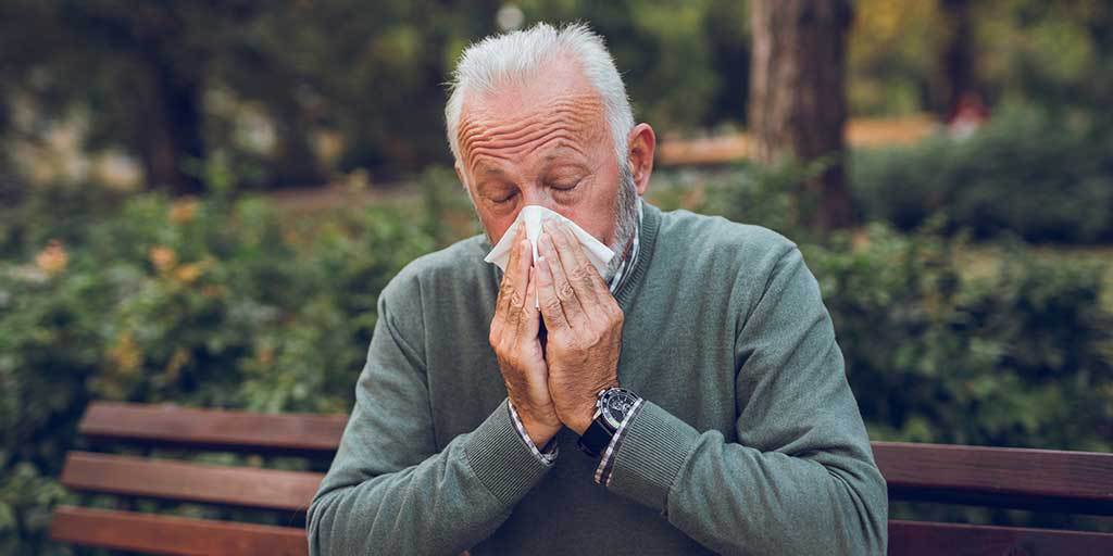 Man sitting on park bench covering nose and mouth with tissue as he blows his nose
