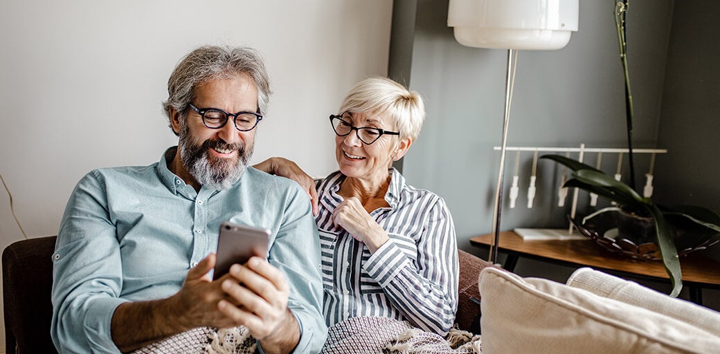 Man and woman comfortably sitting under a blanket together and using mobile phone