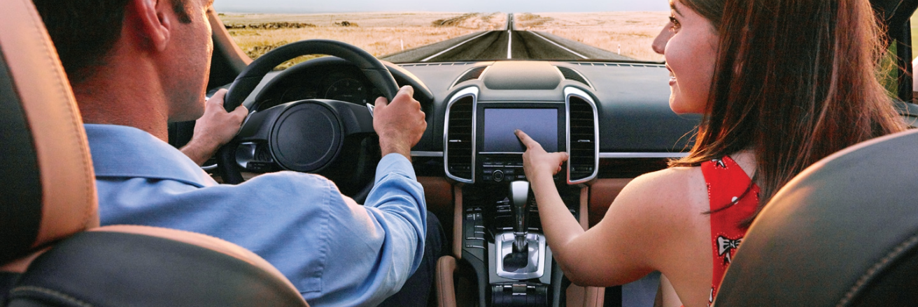 View of a luxury car through the windscreen being driven down a straight road, a man driving with hands on the steering wheel, a woman pointing to the sat nav screen