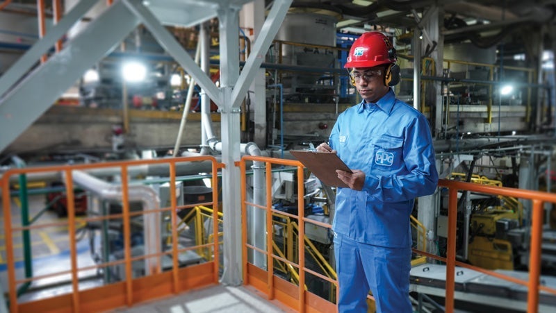 Worker in blue uniform and red hard hat holding a clipboard, inspecting machinery in an industrial plant