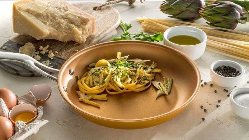 Elegant presentation of pasta with herbs in a copper pan, surrounded by ingredients like parmesan cheese, artichokes, eggs, and olive oil on a marble countertop