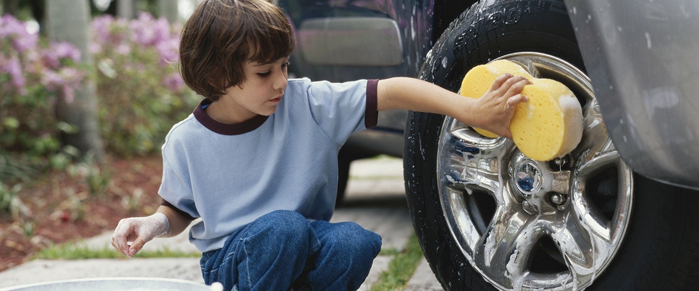 Young child in a blue shirt and jeans kneeling and cleaning a shiny car wheel with a yellow sponge, surrounded by greenery and pink flowers