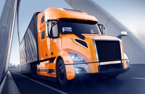 A modern semi tractor trailer is crossing over a bridge. The angle of the photo puts the semi about to pass the viewer on the viewer's right side. The tractor trailer's cab is coated with bright orange finish. The trailer itself is silver. There is no traffic on the highway, and it is a clear, sunny day.