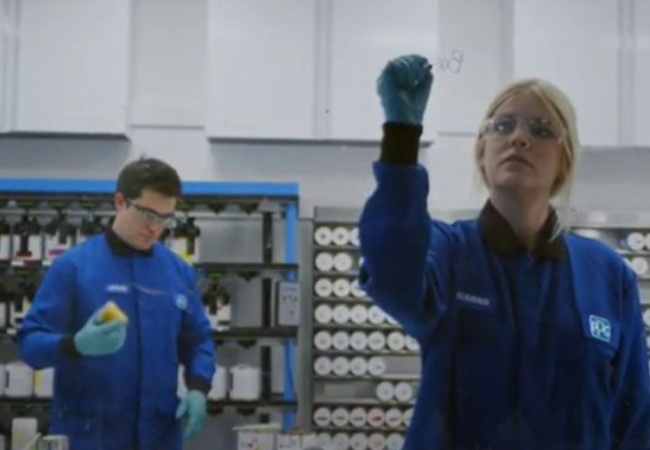 Three workers in blue lab coats engaged in tasks at an industrial paint mixing facility, surrounded by shelves of paint bottles and equipment