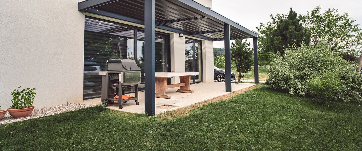 Stylish backyard patio featuring metal cover, wooden furniture, and a barbecue grill, surrounded by green landscaping