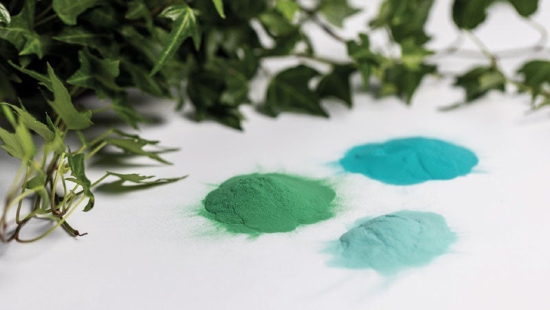 Green and blue pigment powders scattered on a white surface with green plants hanging over them