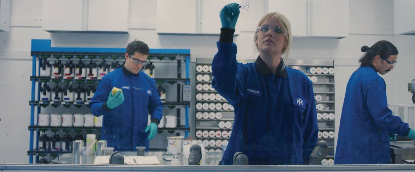 Three workers in blue lab coats engaged in tasks at an industrial paint mixing facility, surrounded by shelves of paint bottles and equipment