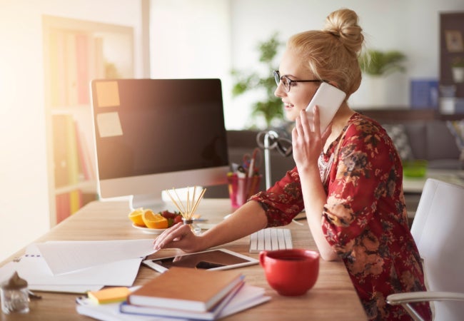 Woman sat at a desk in front of a monitor talking on a white mobile phone